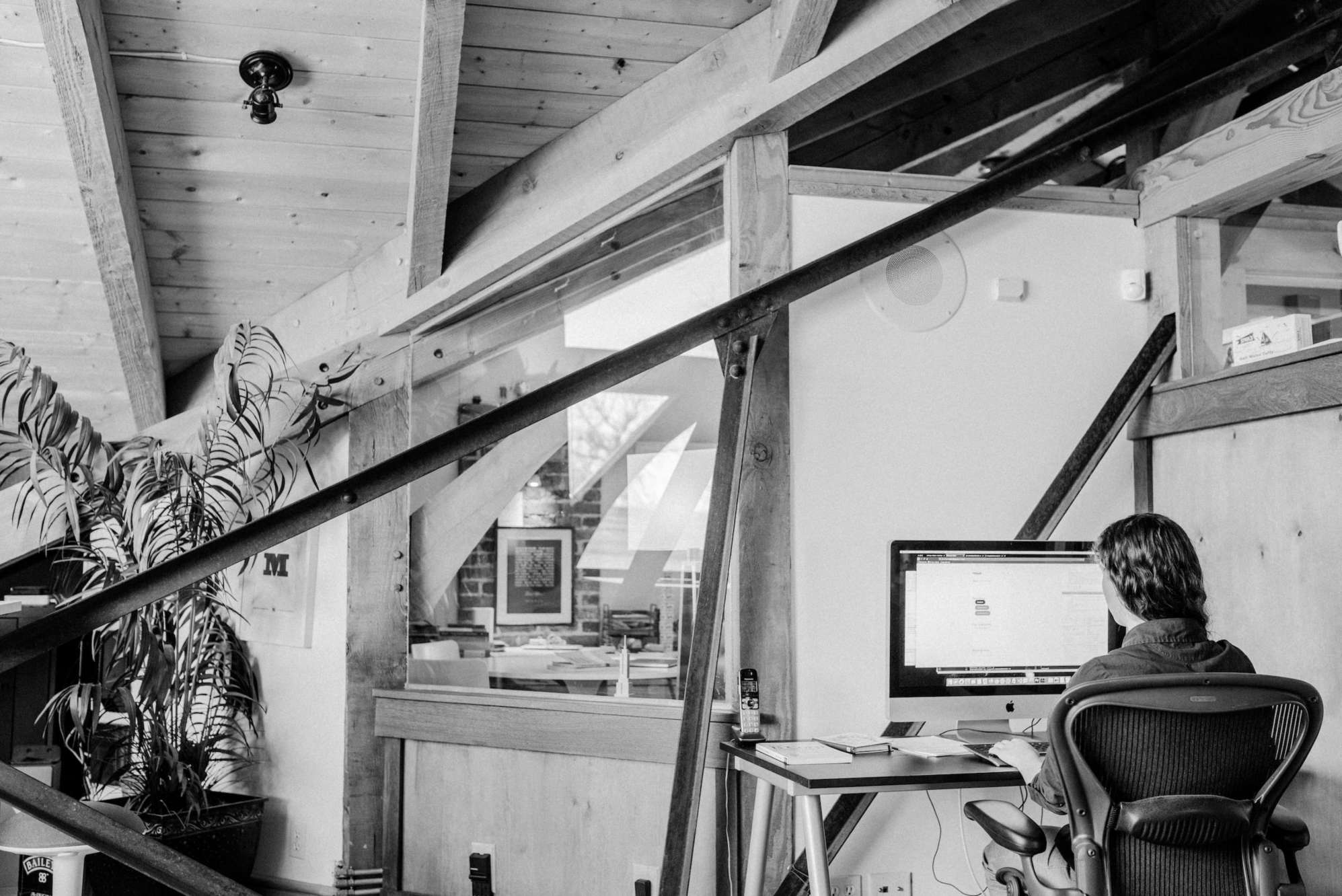 A black and white photo that shows off a corner of the Manoverboard office, with rustic metal and wooden beams.  In the right hand corner, Jennica is seen from the back working at her computer.