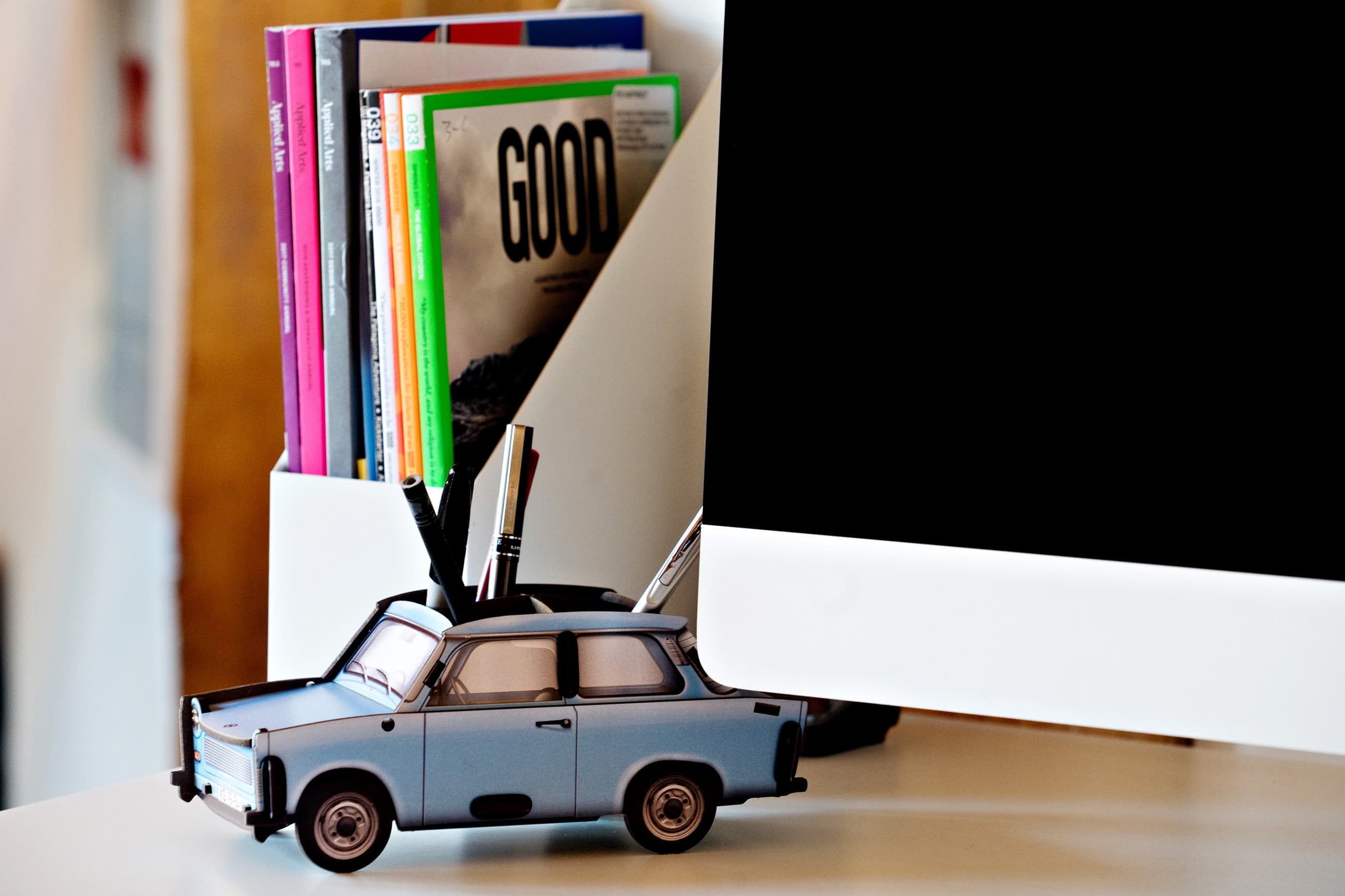 A slice of a manoverboard desk A close up of a corner ofa desk. An iMac screen, a retro toy car, and magazines in folder holders are visible.
