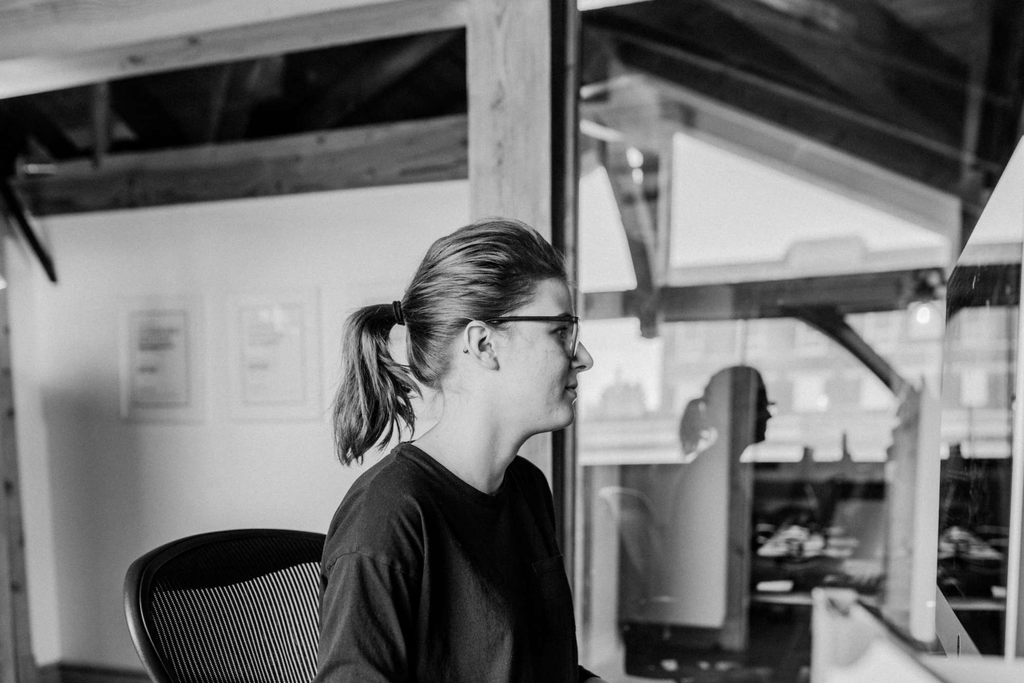 A black and white side profile of Nikki, facing to the right, looking at their computer. Their reflection is seen in the window beside them.