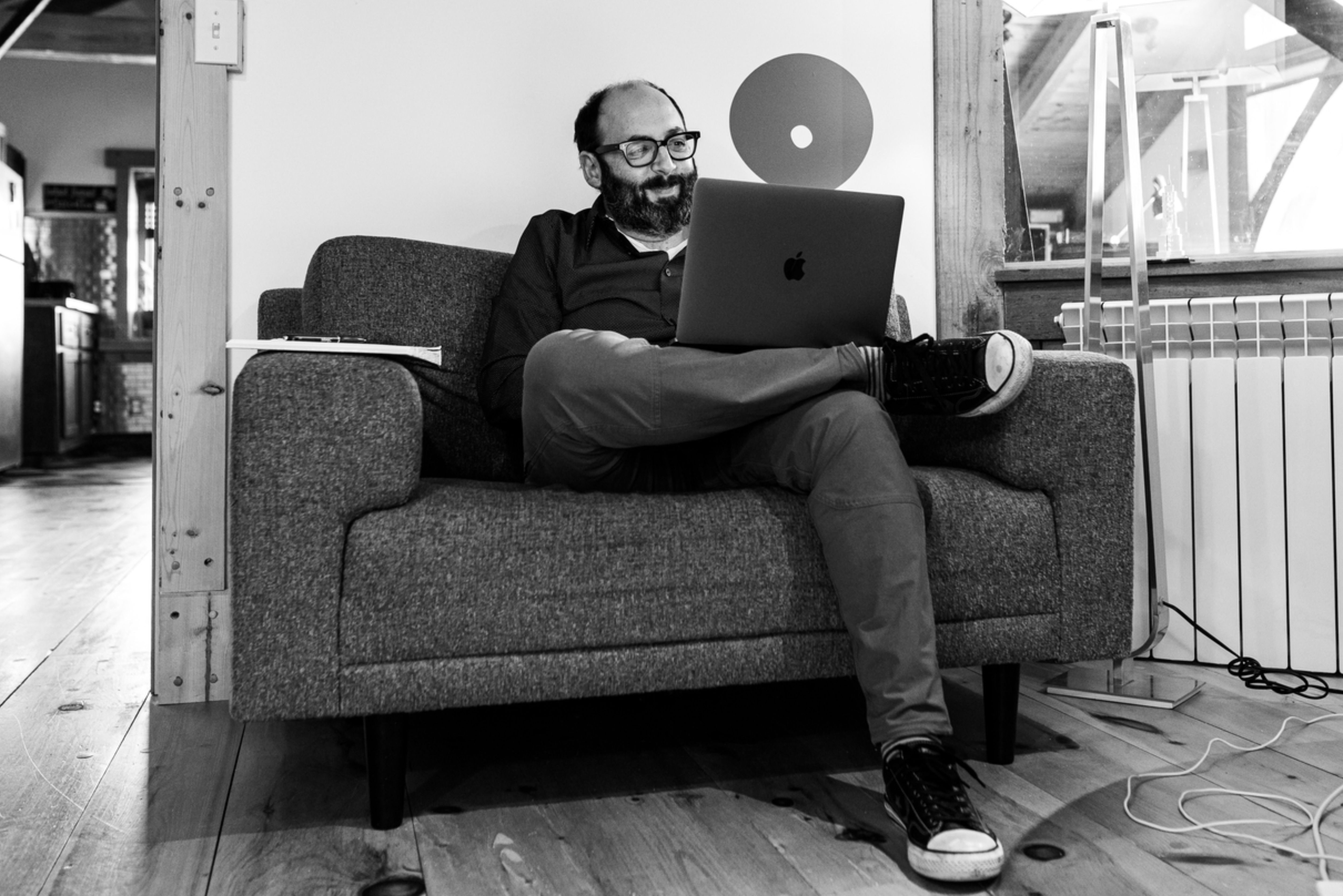 A black and white photo of Andrew sitting on a small couch, smiling and looking at his laptop with his legs crossed.